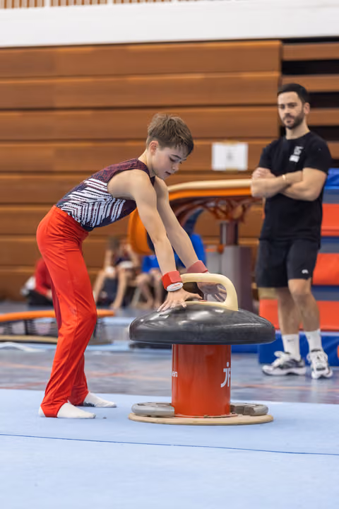 Young gymnast in red pants practicing vault technique on pommel horse while coach observes in gymnasium