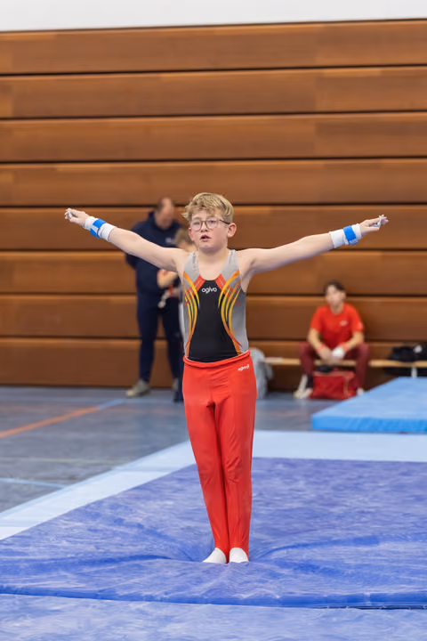 Young gymnast in red pants and black leotard performs floor routine with arms extended, showing focused concentration