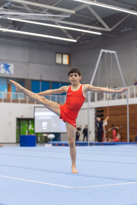 Young gymnast performs vertical split leap on floor exercise mat, arms extended in T-position at indoor training facility