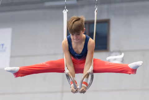Young gymnast performs horizontal split on still rings, demonstrating strength and flexibility in red pants and blue leotard