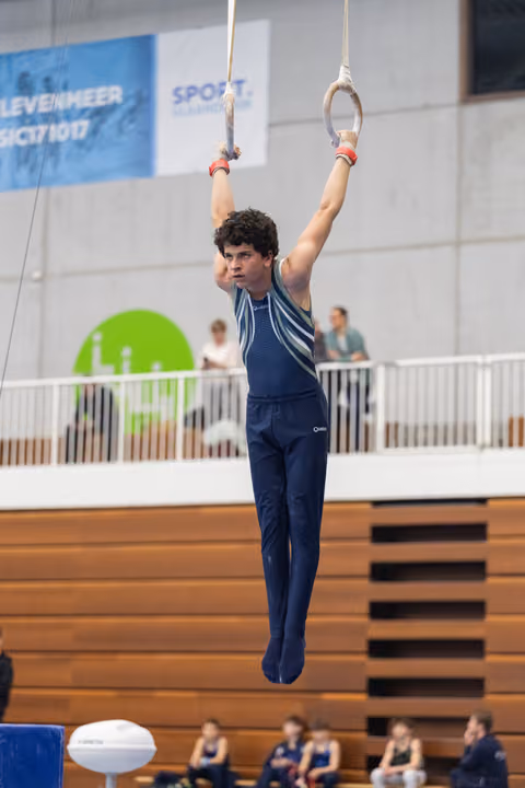 Young male gymnast in dark blue outfit hangs from still rings with focused expression during indoor gymnastics event
