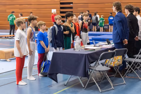 Group of young male gymnasts in sportswear line up at a registration table, engaging with officials in an indoor facility