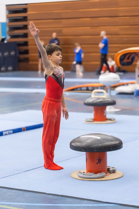 Young gymnast in red uniform stands in salute position beside pommel horse, displaying perfect form during training session