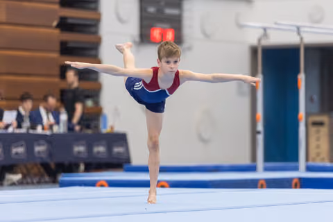 Young gymnast performs an arabesque balance during floor routine, arms extended gracefully at indoor gymnastics venue