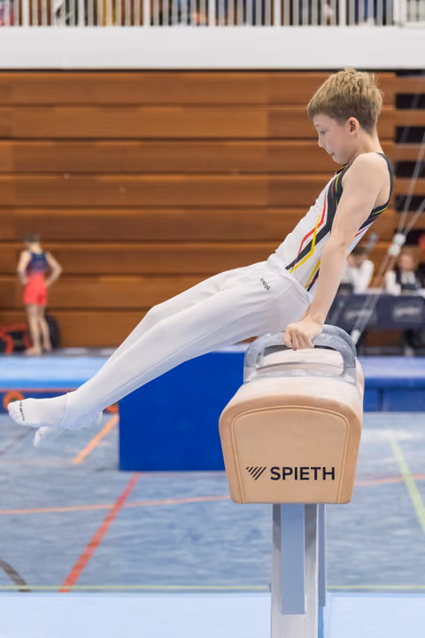 Young gymnast in white uniform executes a pommel horse routine with extended legs during indoor gymnastics meet