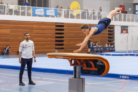 Male gymnast performs vault over Spieth table with extended body position while coach observes from the floor