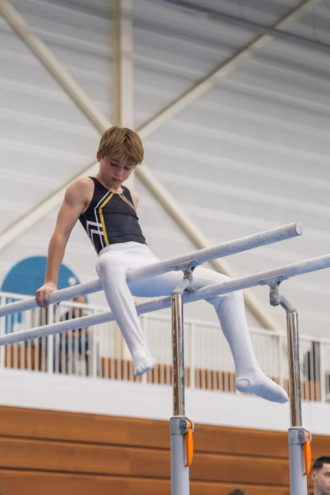 Young gymnast in navy leotard performs on parallel bars, legs extended forward in seated position at indoor gymnastics facility