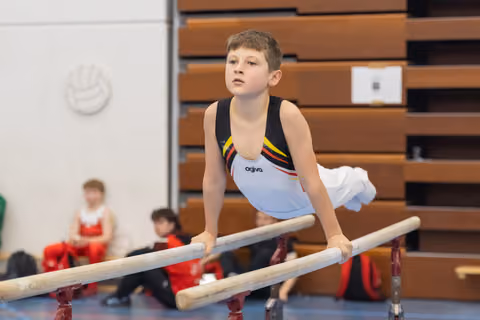 Young gymnast performs on parallel bars with intense concentration, while teammates in red wait in background