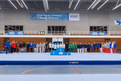 Multiple gymnastics teams lined up on award podium and floor exercise area during medal ceremony in sports hall