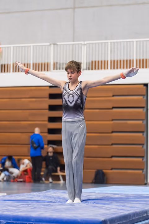 Young male gymnast stands with arms extended horizontally on floor mat, displaying control and form during routine