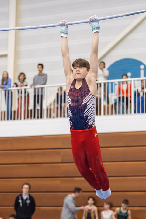 Young gymnast in sparkly leotard and red pants hangs from horizontal bar, arms extended overhead, spectators watching from balcony