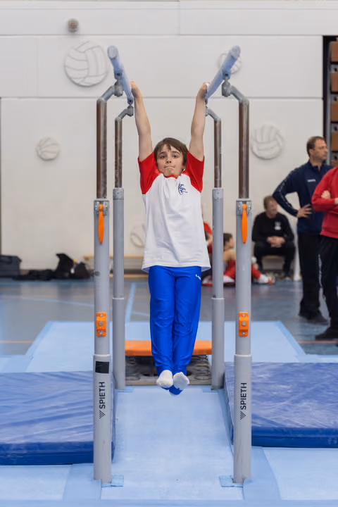 Young gymnast in red and white uniform hangs from parallel bars with arms extended upward during training session