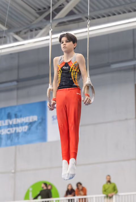 Gymnast performing still rings routine in red pants and flame-design leotard, demonstrating intense focus and control