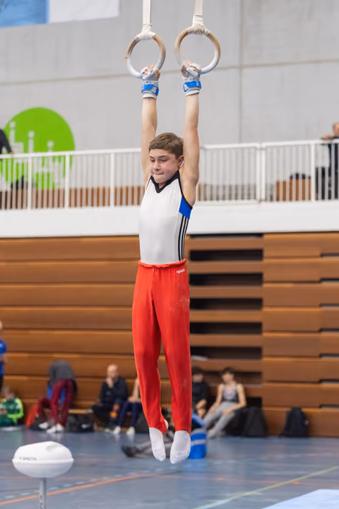 Young gymnast performing a still rings routine, arms extended overhead, wearing red pants and white top in an indoor gym