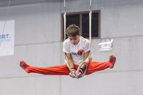 Young gymnast performs L-sit straddle hold on rings, displaying concentration and strength in training facility
