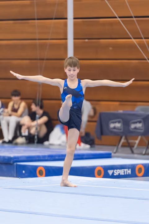 Young gymnast performing a standing balance pose with arms extended and one leg raised during floor exercise routine