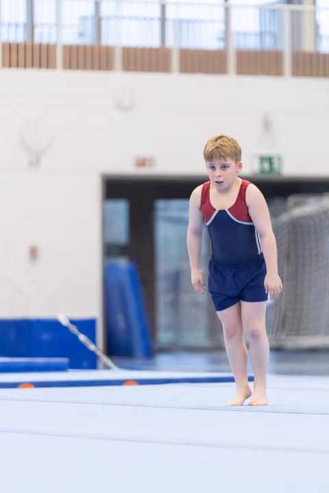 Young gymnast in navy and red leotard walks barefoot on floor mat, focused expression, training facility with blue equipment