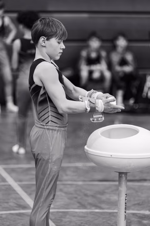 Young male gymnast chalks his hands before pommel horse routine, focused and preparing in gym with blurred figures behind