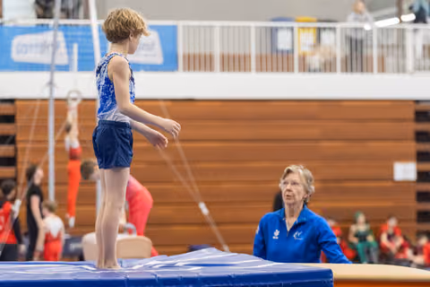 Young gymnast stands on vault apparatus while female coach in blue jacket observes from below during training session