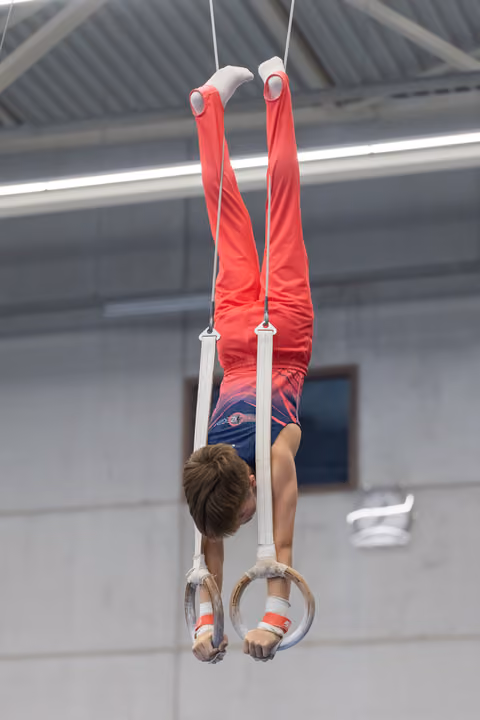 Young gymnast performing an inverted hang on rings, legs extended vertically upward in red uniform during training
