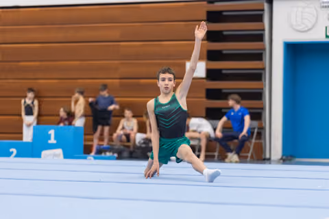 Male gymnast performs lunge pose with arm raised during floor exercise routine in gymnasium