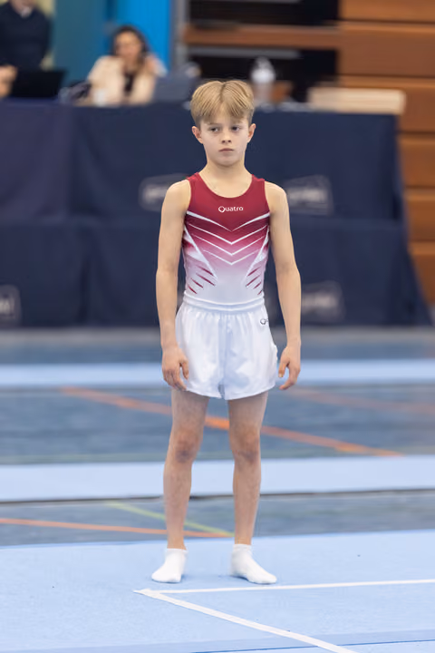 Young gymnast in burgundy and white leotard stands at attention on blue floor mat, focused and ready to perform