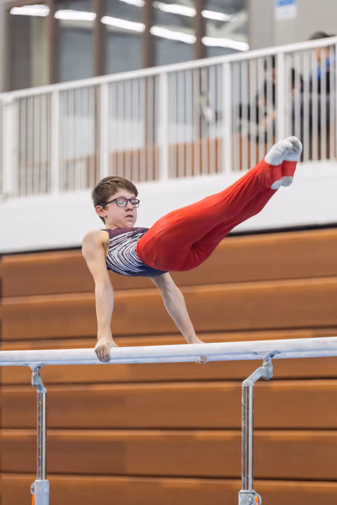 Young gymnast in glasses performing L-sit hold on parallel bars, demonstrating strength and focus in training facility