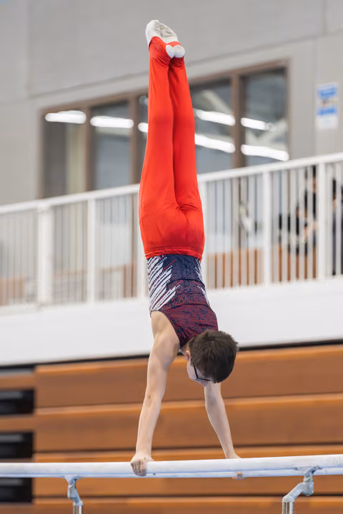 Young gymnast performs vertical handstand on horizontal bar in gymnasium, displaying strength and balance in red outfit