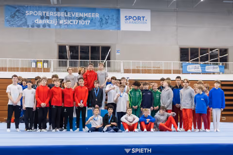 Large group of young male gymnasts in colorful training gear posing together on a blue mat in an indoor gymnasium