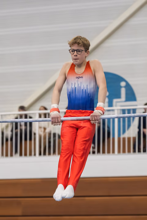 Young gymnast in red and blue gradient uniform concentrates while performing on horizontal bar, wearing glasses