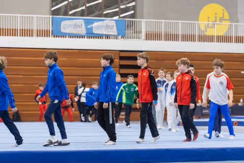 Young gymnasts in team tracksuits march in formation across a blue floor mat during opening ceremony in a gymnasium