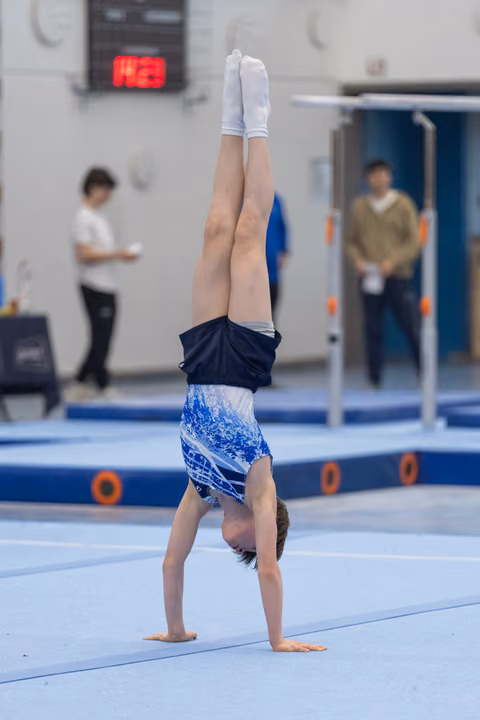 Young gymnast executes a handstand on blue mat, legs straight and pointed, while coaches observe in background