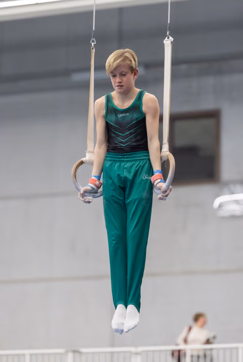 Young gymnast with blonde hair holds steady on still rings, focused expression, wearing green competition uniform in training hall