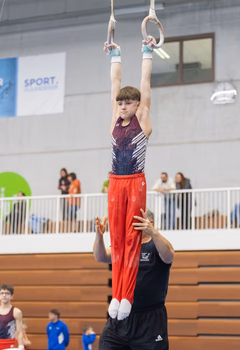 Young gymnast in red pants hangs from rings with focused expression while coach spots from below in indoor gym