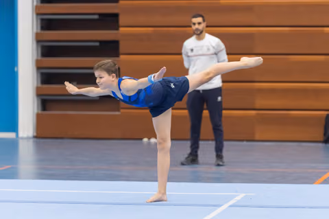 Young gymnast in blue leotard executes an arabesque balance on floor exercise while coach observes from background