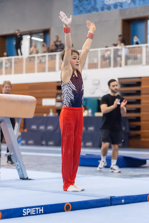 Young gymnast in red pants and patterned leotard stands at attention with arms raised, preparing for balance beam routine