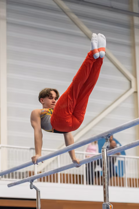 Male gymnast demonstrates an L-sit position on parallel bars, wearing red pants and grey top, indoors training facility