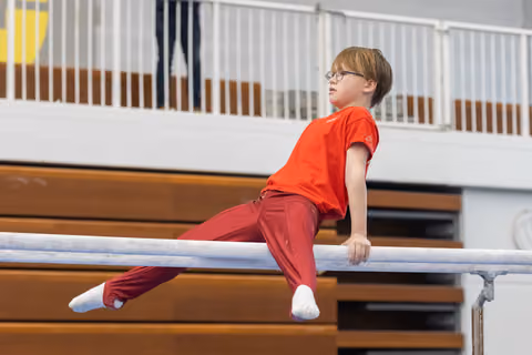 Young gymnast with glasses performing a support hold on parallel bars, focused expression, wearing red outfit in gymnasium