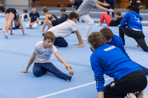 Young gymnast in white shirt stretches on blue mat while coach in blue jacket provides hands-on guidance and support