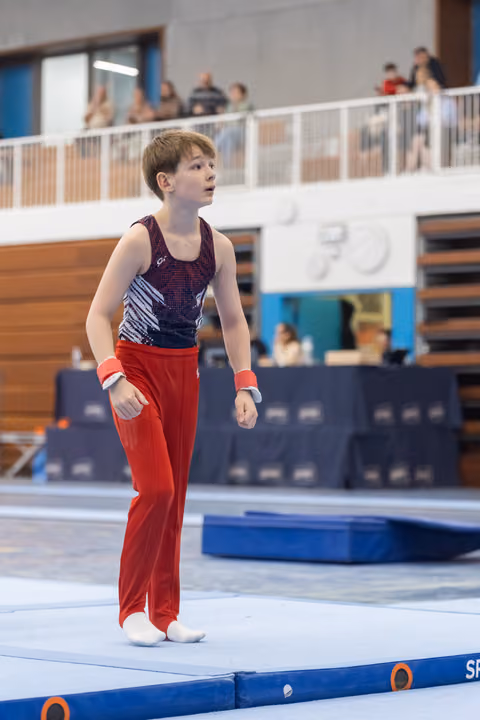 Young gymnast in purple leotard and red pants stands thoughtfully on floor mat, gazing upward during indoor gymnastics event