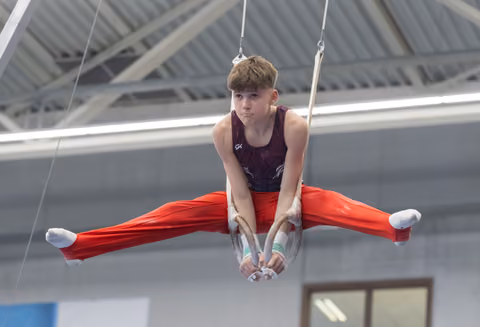 Male gymnast performs straddle position on still rings, displaying strength and control in indoor training facility