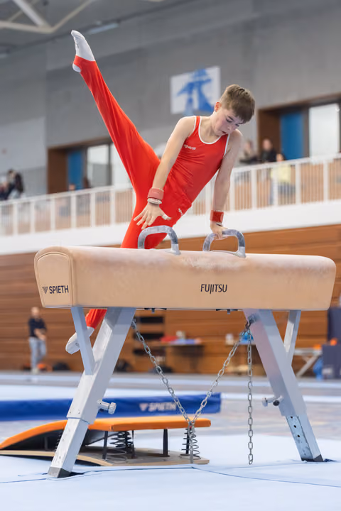 Young gymnast in red leotard executes a split-leg position on pommel horse, demonstrating strength and control in training facility