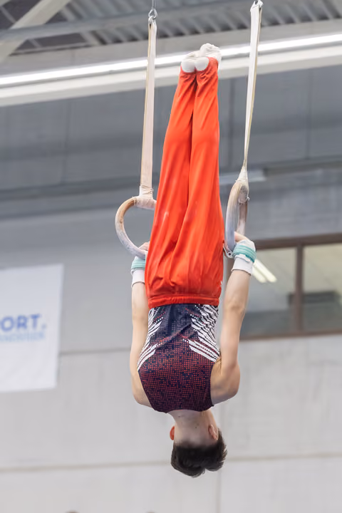 Young gymnast performing an inverted hang on still rings, wearing red top and patterned shorts in indoor training facility