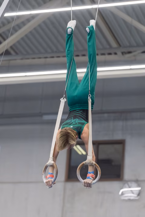 Young gymnast performs vertical hang on rings, displaying strength and control while wearing teal uniform in indoor facility