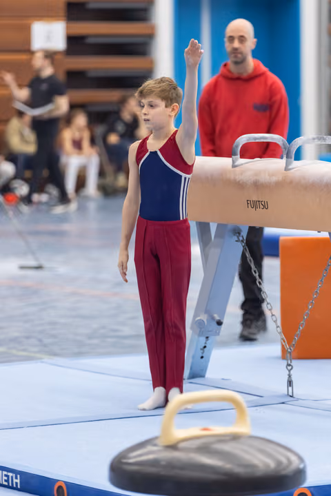 Young gymnast stands beside pommel horse with raised hand, focused expression, while coach observes in background