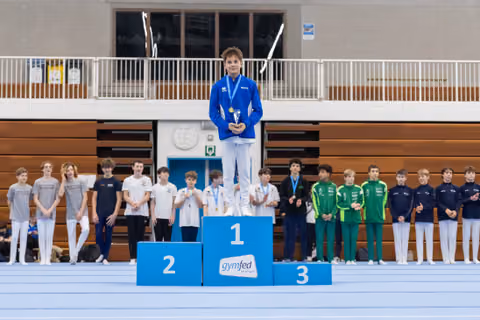 Young gymnast in blue tracksuit stands atop first place podium holding medal, with competing teams lined up behind