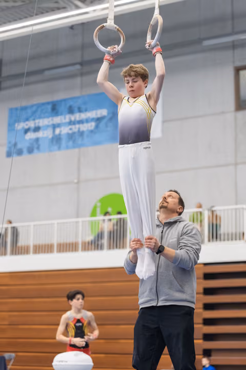 Young gymnast holds onto rings while coach supports him from below during training at indoor gymnasium