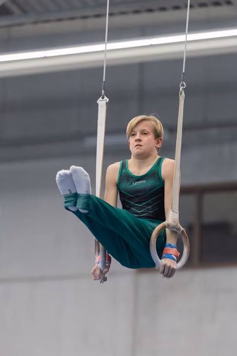 Young gymnast in green leotard performing L-sit position on rings, displaying intense focus and strength during routine