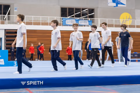 Six young male gymnasts in white team uniforms march in formation across a blue Spieth floor mat during a team presentation