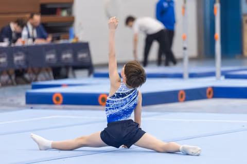 Young gymnast performs floor split routine in blue sequined leotard with arm extended upward during training session